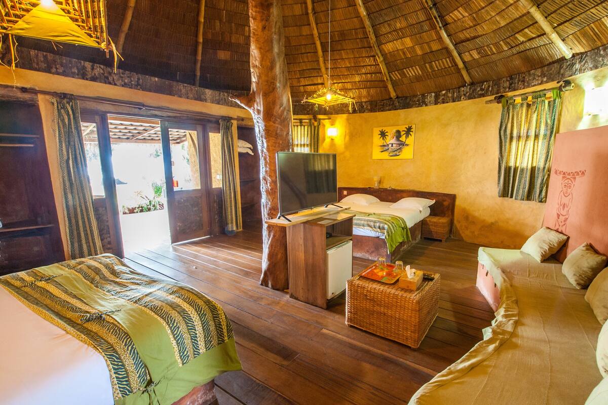 Quarto com piso de madeira, cama com colcha estampada em verde e amarelo, sofá com almofadas, televisão sobre um suporte de madeira e parede com pintura tropical.