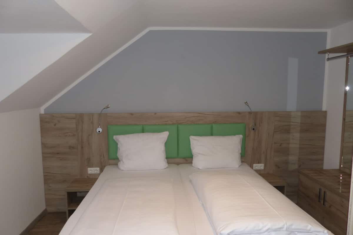 A small, single-room hotel bedroom with a sloped ceiling, a wooden headboard, two beds with white bedding, and a bedside table.