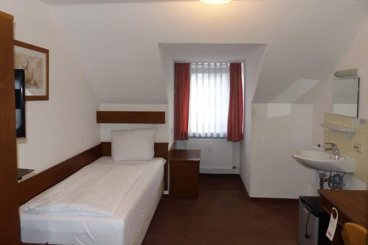 A hotel room with a single bed, a bedside table, a desk with a chair, a window with curtains, a bathroom with a sink and mirror, and a framed picture on the wall.