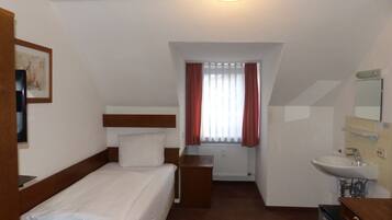 Habitación de hotel con una cama individual, una mesita de noche, un escritorio con silla, una ventana con cortinas, un baño con lavamanos y espejo, y un cuadro en la pared.