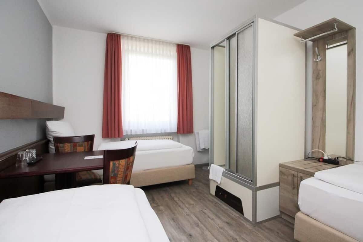 A hotel room with a bed, a desk with a chair, a small table with a glass, a mirror, and a shower area.
