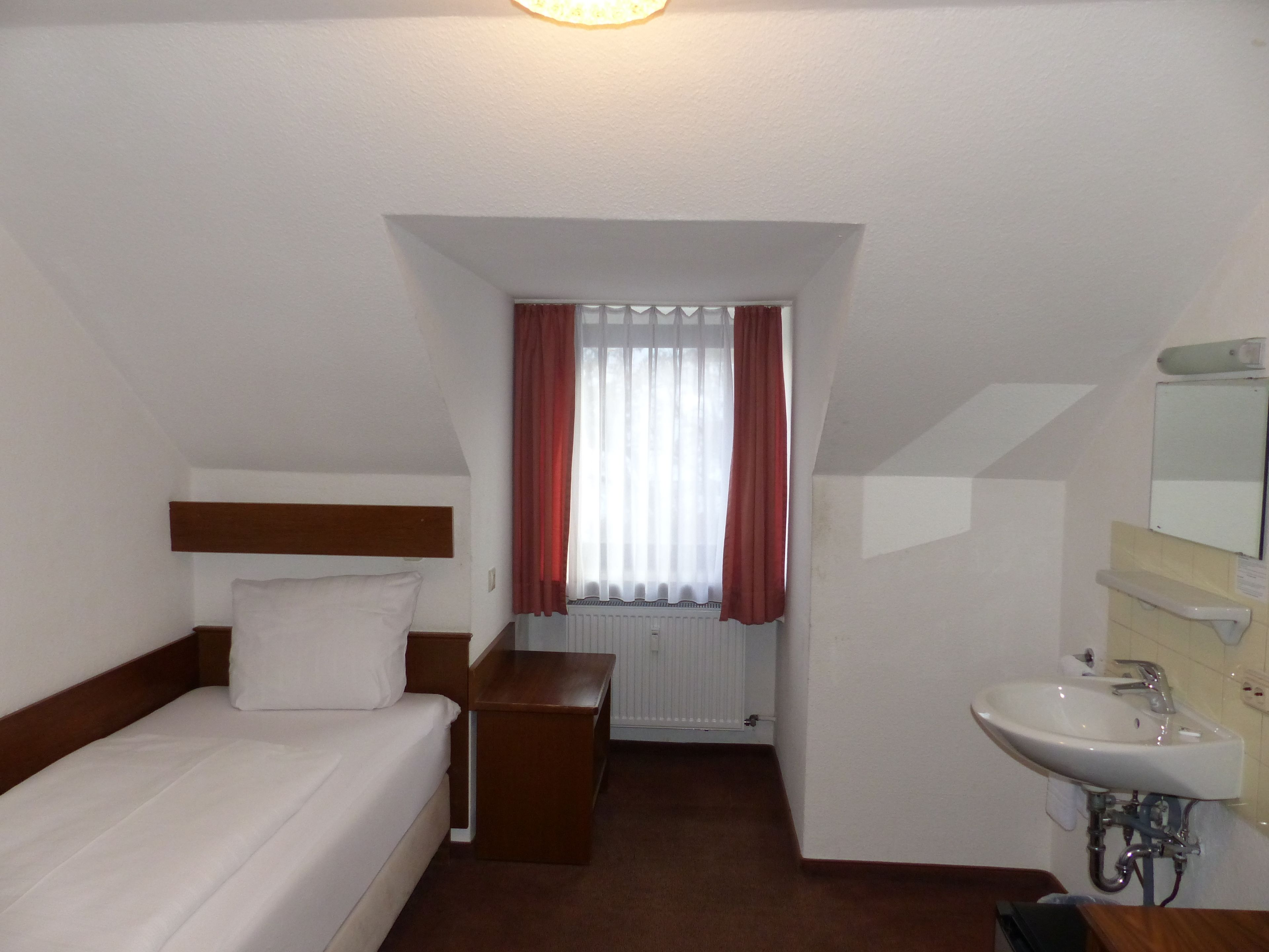 A hotel room with a single bed, a wooden nightstand, a small table, a window with red curtains, a radiator, and a bathroom with a sink and mirror.