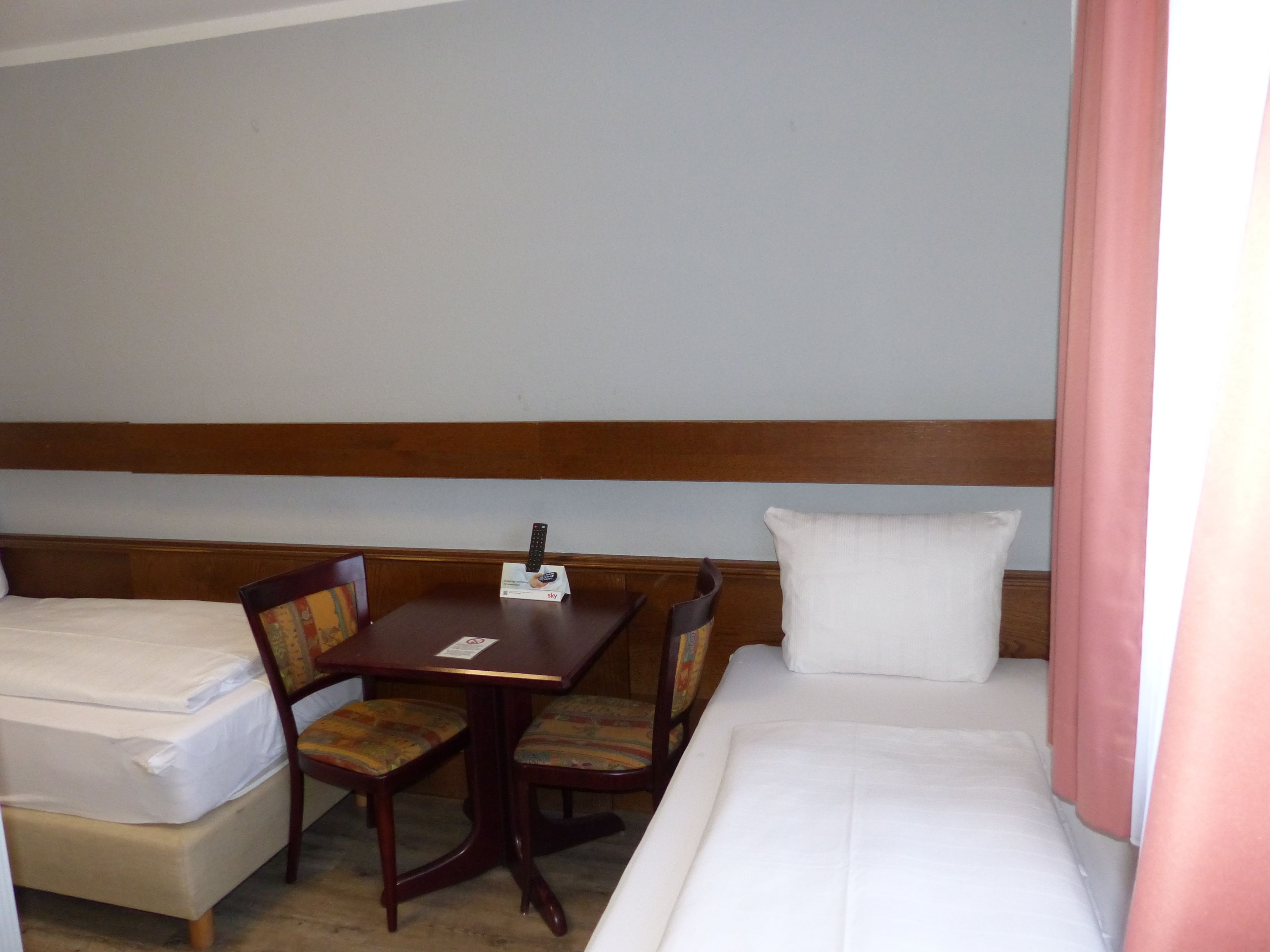 Habitación de hotel con dos camas individuales, una mesa de madera con un control remoto y dos sillones tapizados.