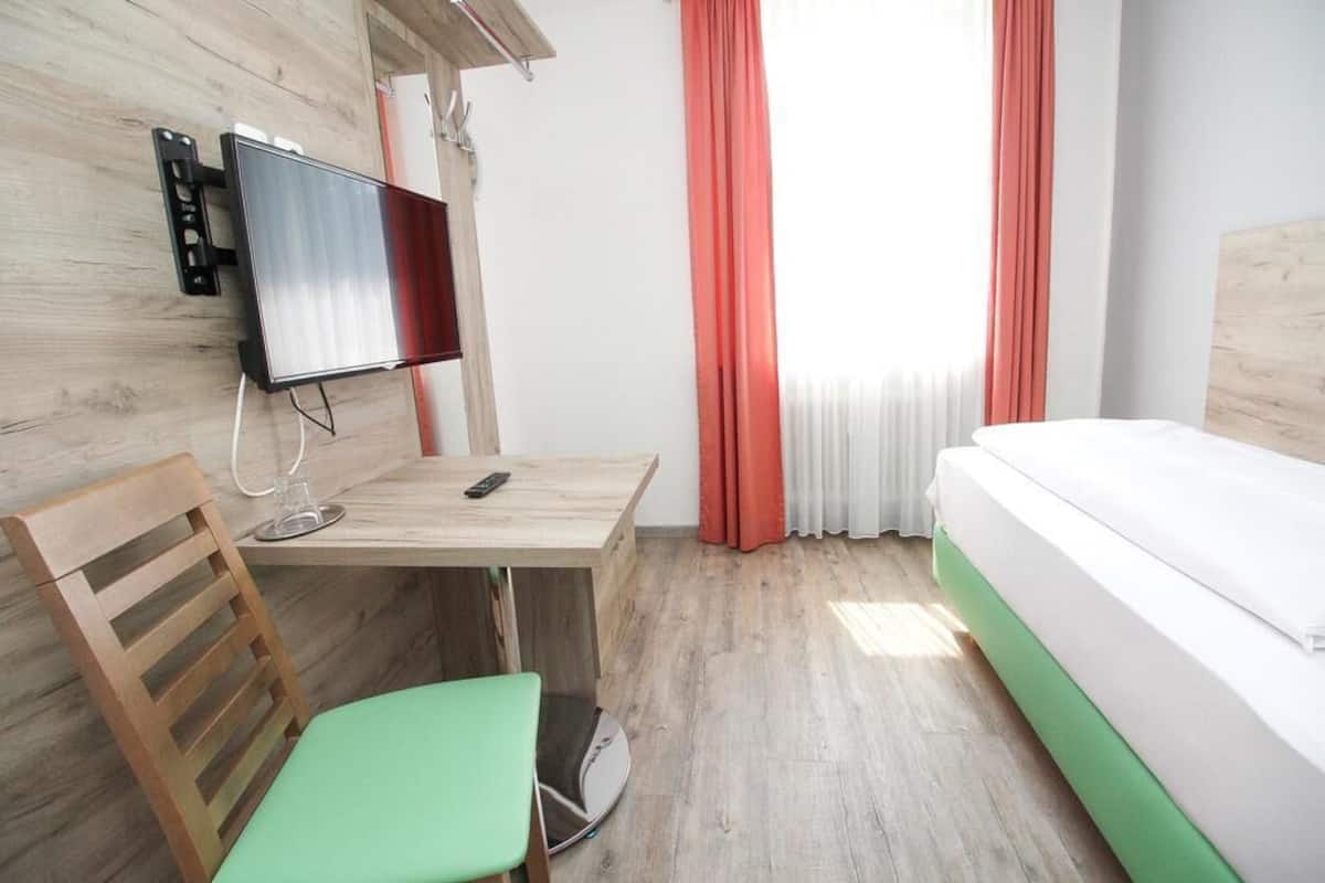 A hotel room with a wooden desk, a green chair, a bed with a green mattress, a TV mounted on the wall, and red curtains.
