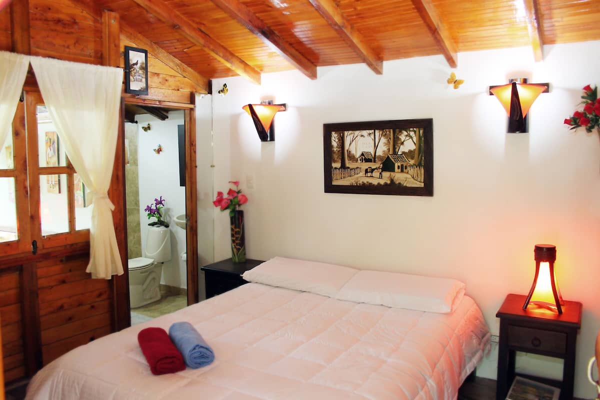 Une chambre à coucher avec un lit, une table de chevet avec une lampe, un tableau au mur et une salle de bain visible à travers une porte ouverte.