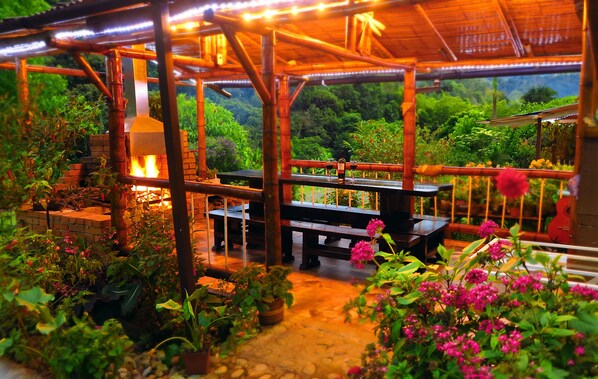 Enjoy meals under a rustic bamboo structure surrounded by lush greenery and a fire pit.