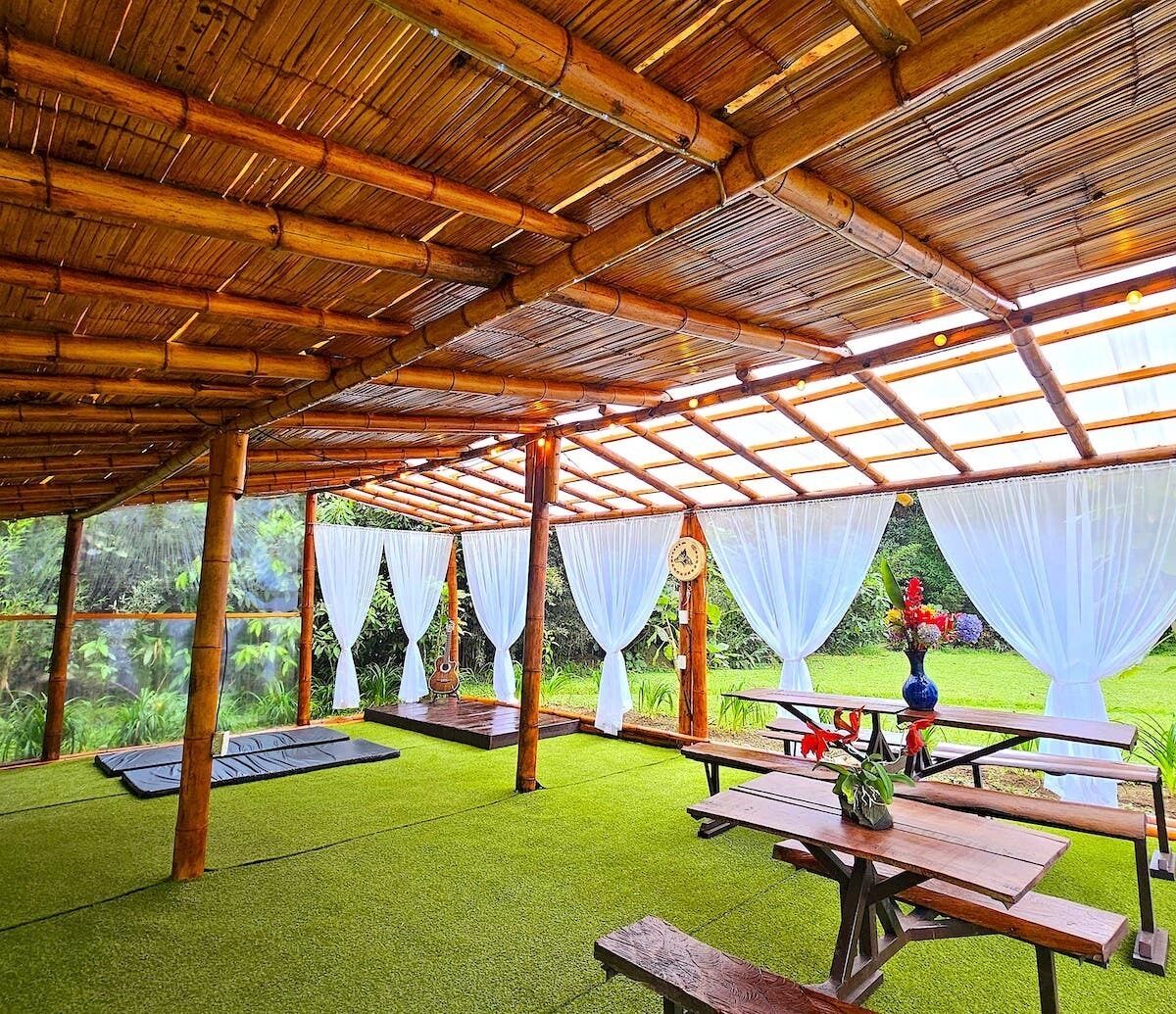 Un espacio exterior techado con vigas de bambú y cortinas blancas, que incluye mesas de picnic y un reloj en la pared.