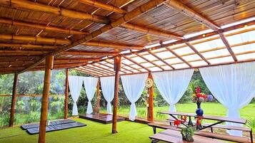 Un espacio exterior techado con vigas de bambú y cortinas blancas, que incluye mesas de picnic y un reloj en la pared.