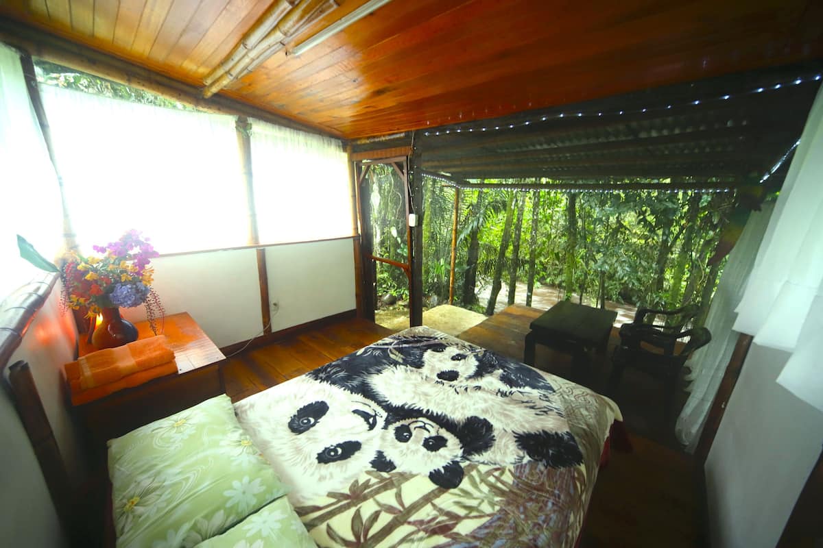 Une chambre à coucher avec un lit, une table de chevet ornée d’un vase de fleurs et une vue sur un jardin luxuriant, visible à travers une porte vitrée.