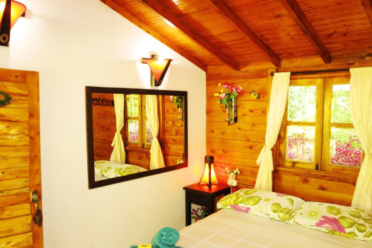 Une chambre dans un chalet en bois, avec un lit, un miroir, une table de chevet avec une lampe et une fenêtre avec des rideaux.