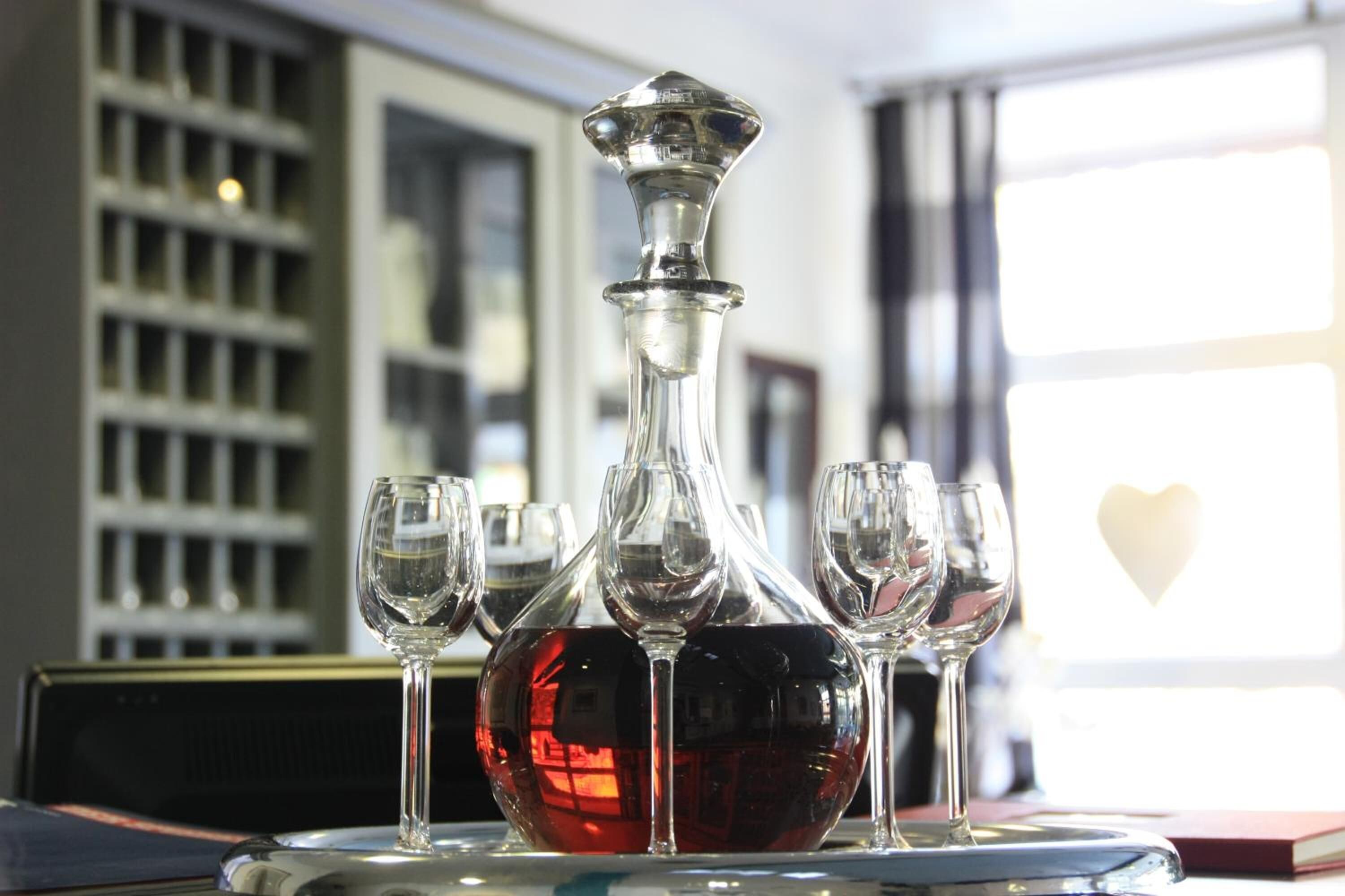 A decanter with wine glasses on a table, with a bookshelf and a window in the background.
