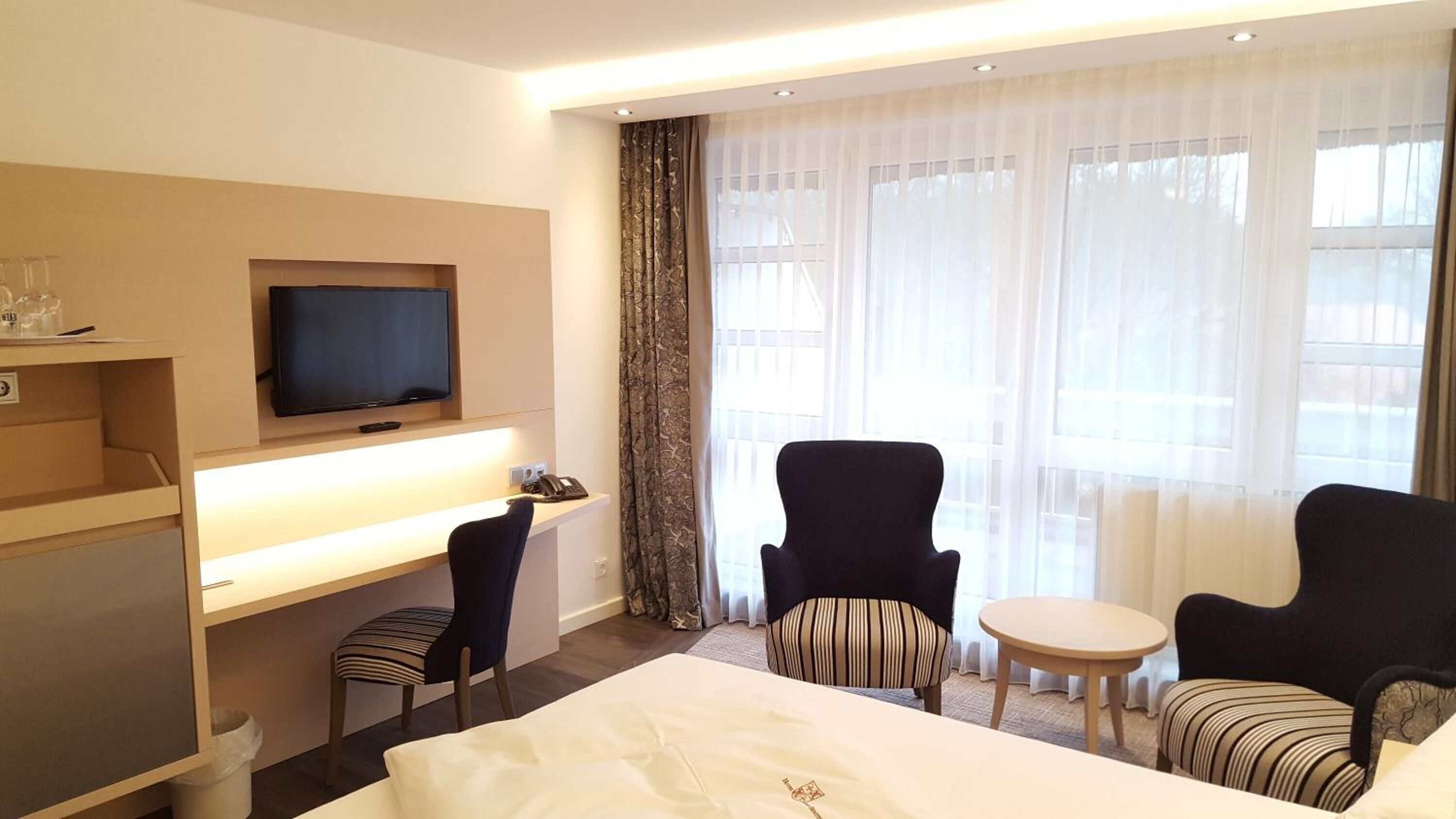 A hotel room with a bed, a desk with a TV, two armchairs, a small table, and a window with sheer curtains.