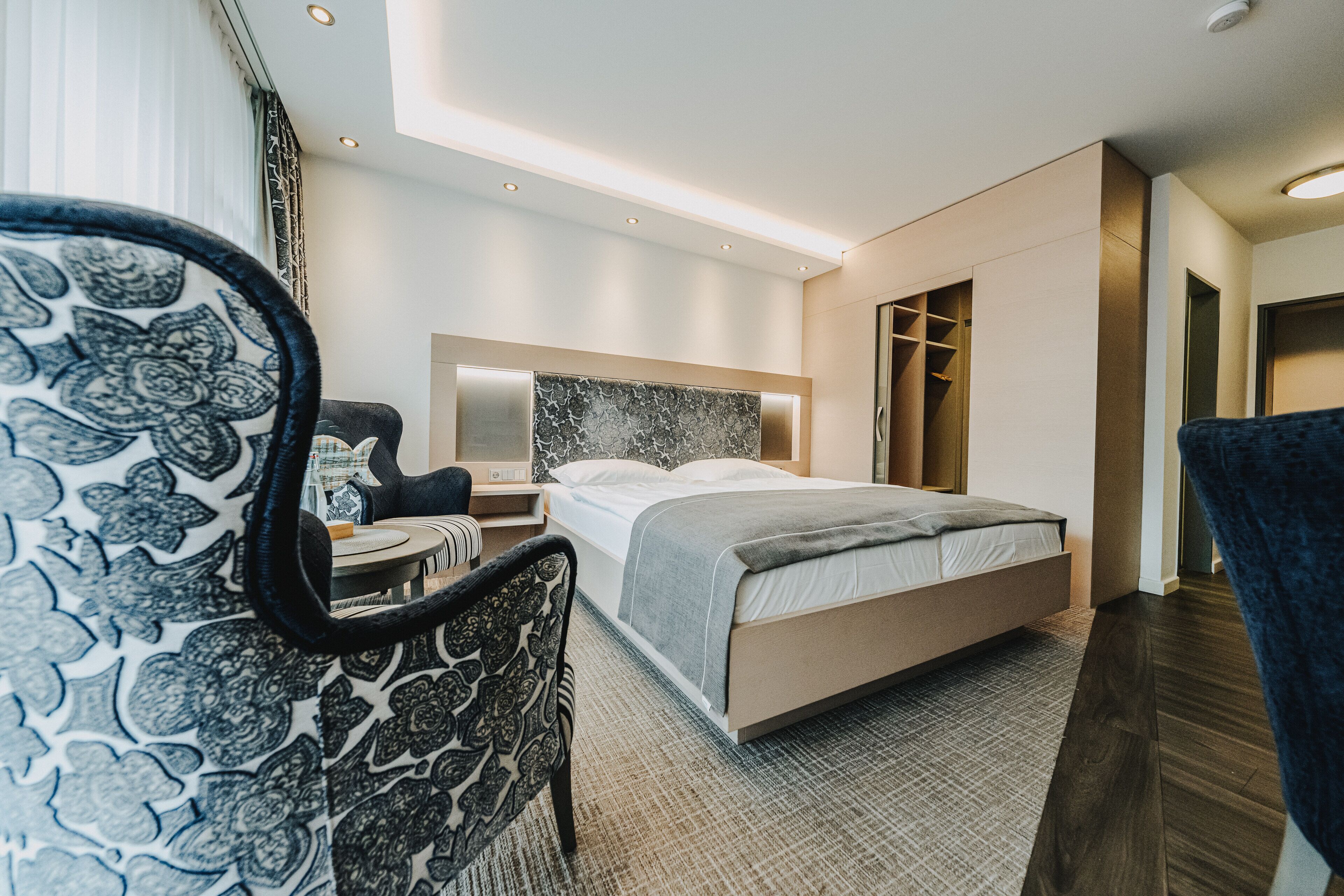 A modern hotel room with a bed, bedside table, chair, and built-in wardrobe.