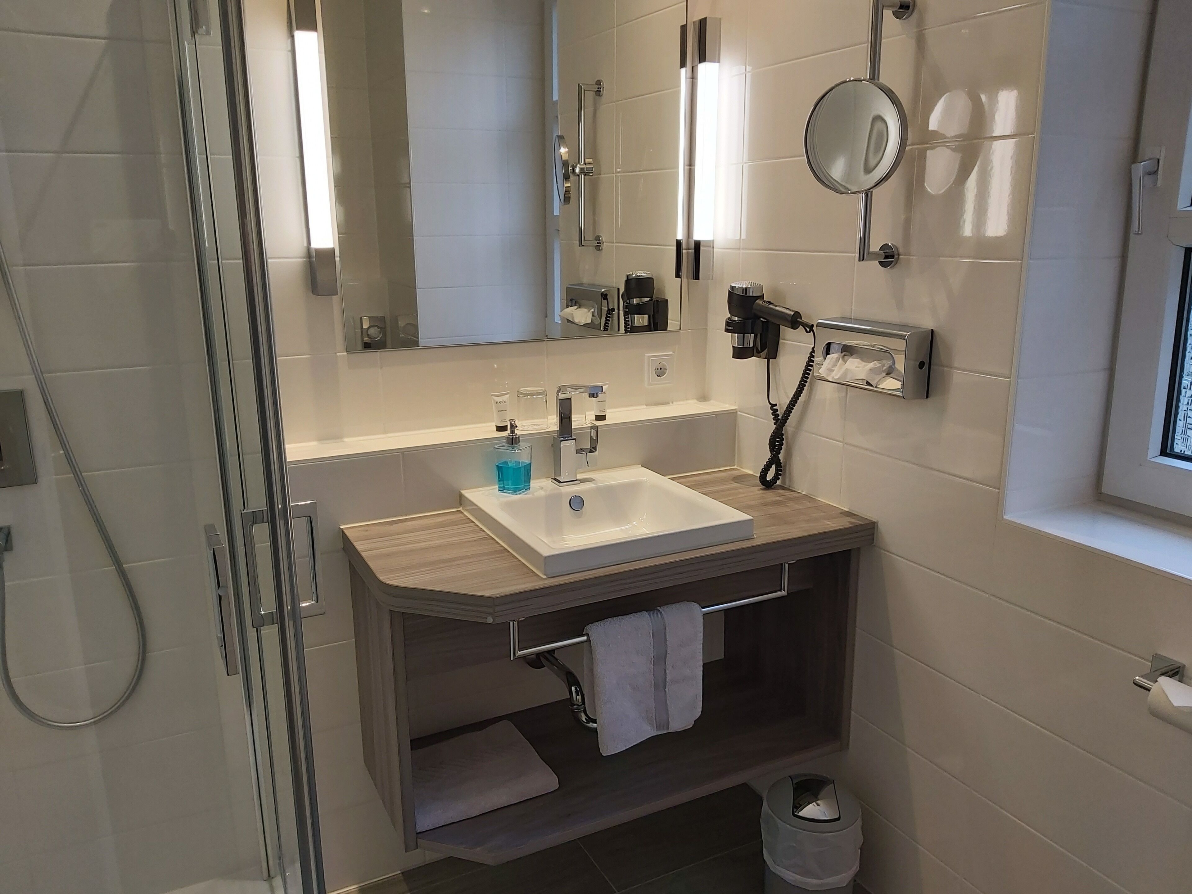A modern bathroom with a glass shower enclosure, a white sink with a wooden countertop, and a mirror with a round frame.