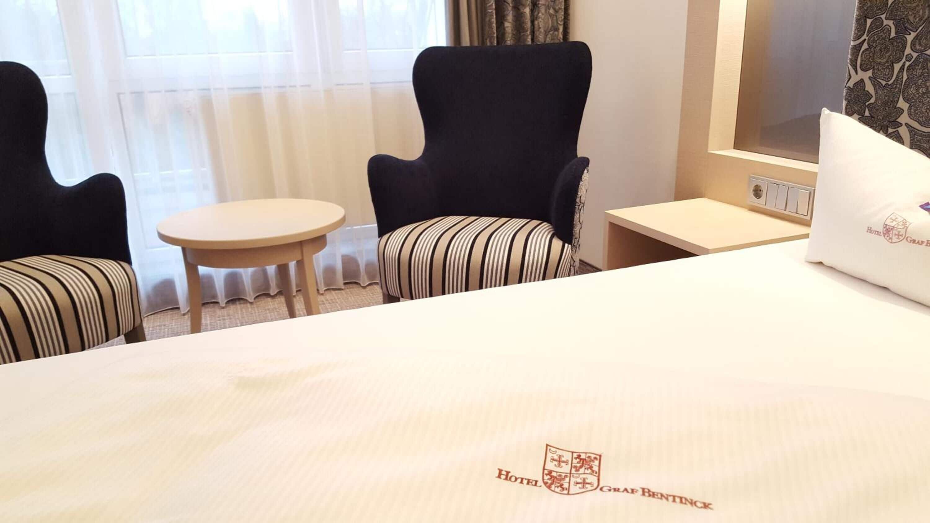 Una habitación de hotel con una cama, dos sillones, una mesita redonda y una mesita de noche con una lámpara.