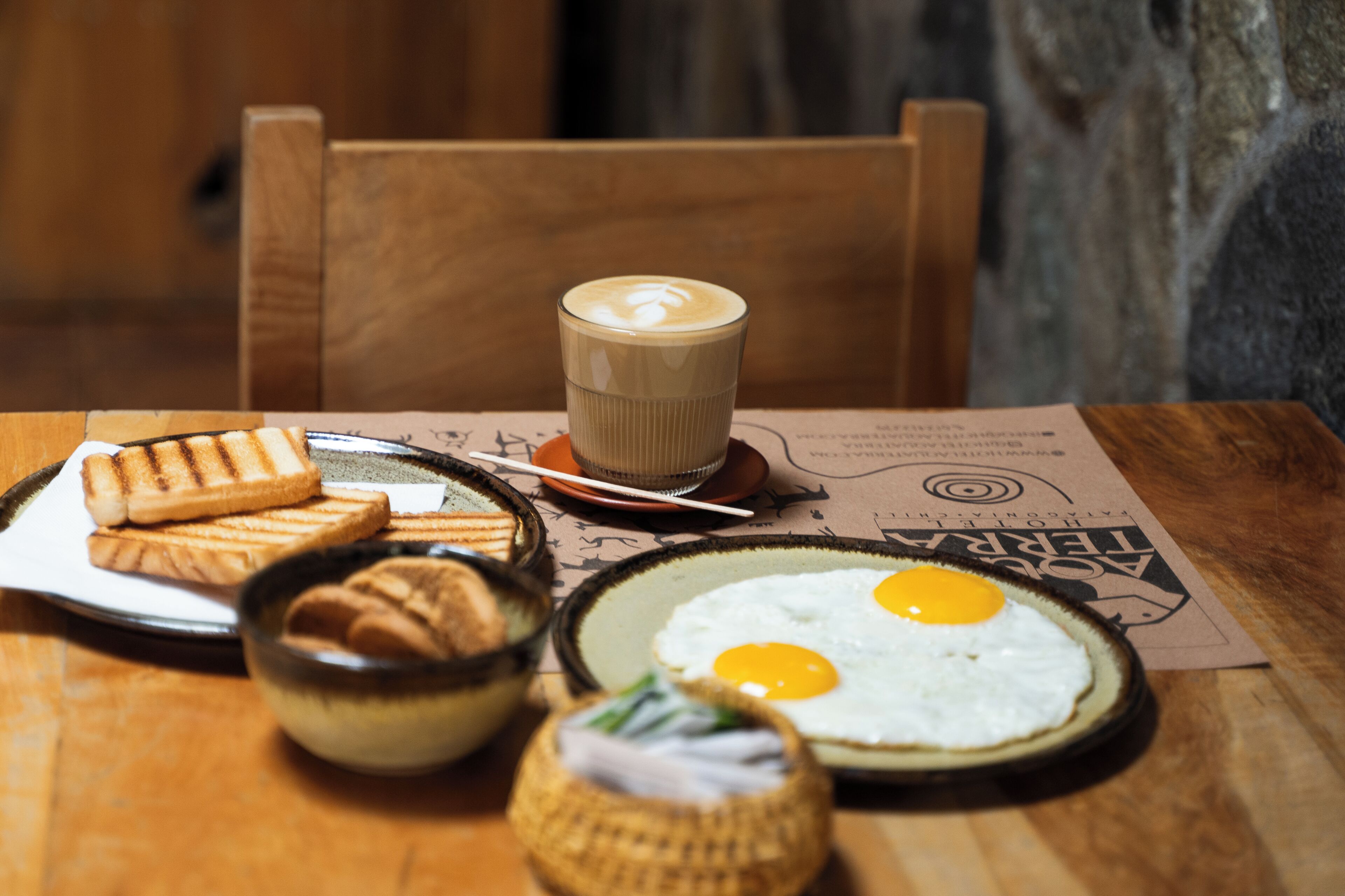Ein Holztisch mit einer Platte Grillbrot, einer Schüssel eingelegtes Gemüse, einer Platte mit zwei Spiegeleiern und einem Kaffeeglas.