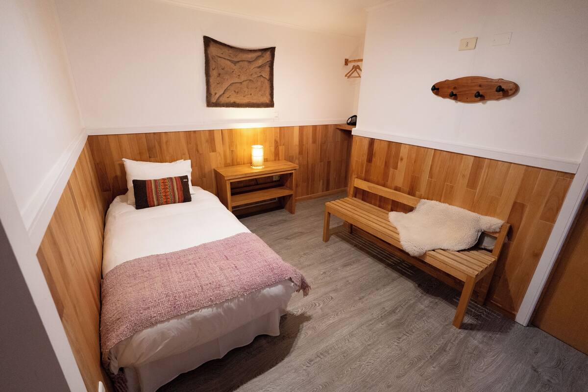 Una habitación pequeña y acogedora con una cama, un banco, una mesita de noche con una lámpara y un estante de madera en la pared.