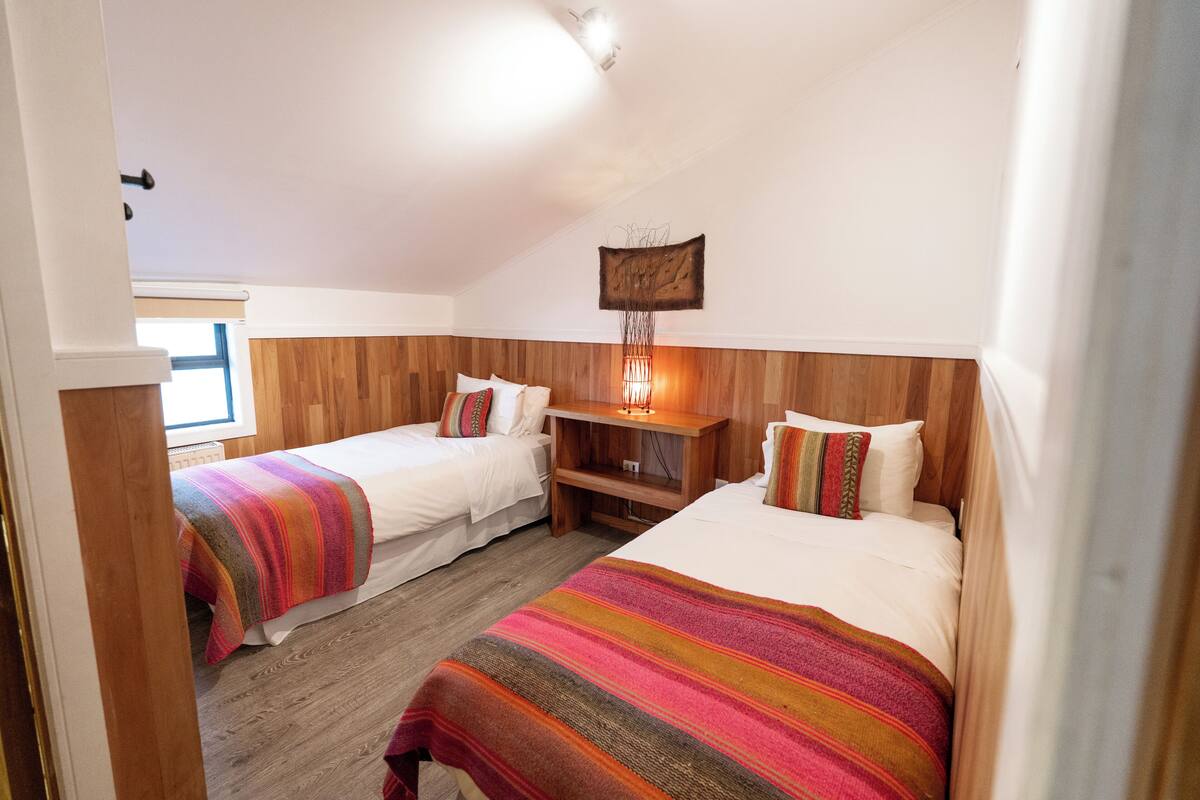 Una habitación con dos camas, cabeceros de madera, una mesita de noche, una lámpara fijada a la pared y una ventana.