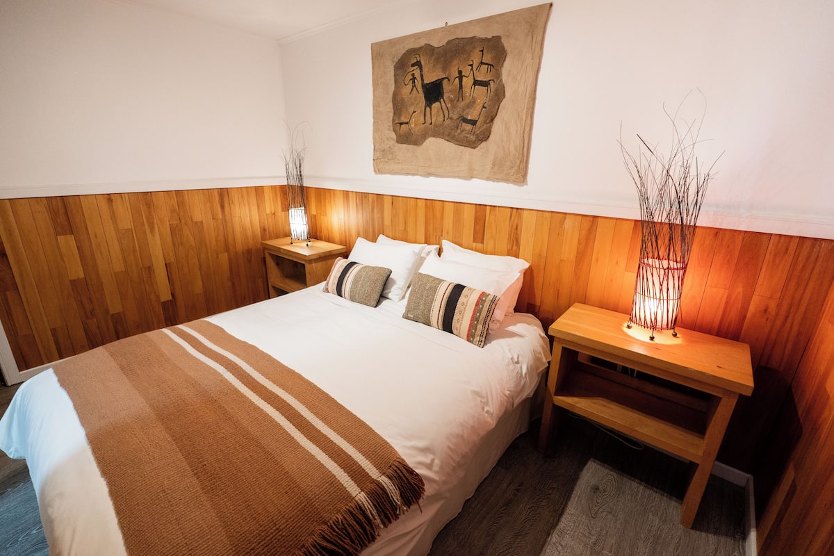 Un dormitorio con cabecera de madera, cama con ropa de cama blanca y un plaid a rayas, dos mesitas de noche con lámparas, y un tapiz colgado en la pared.