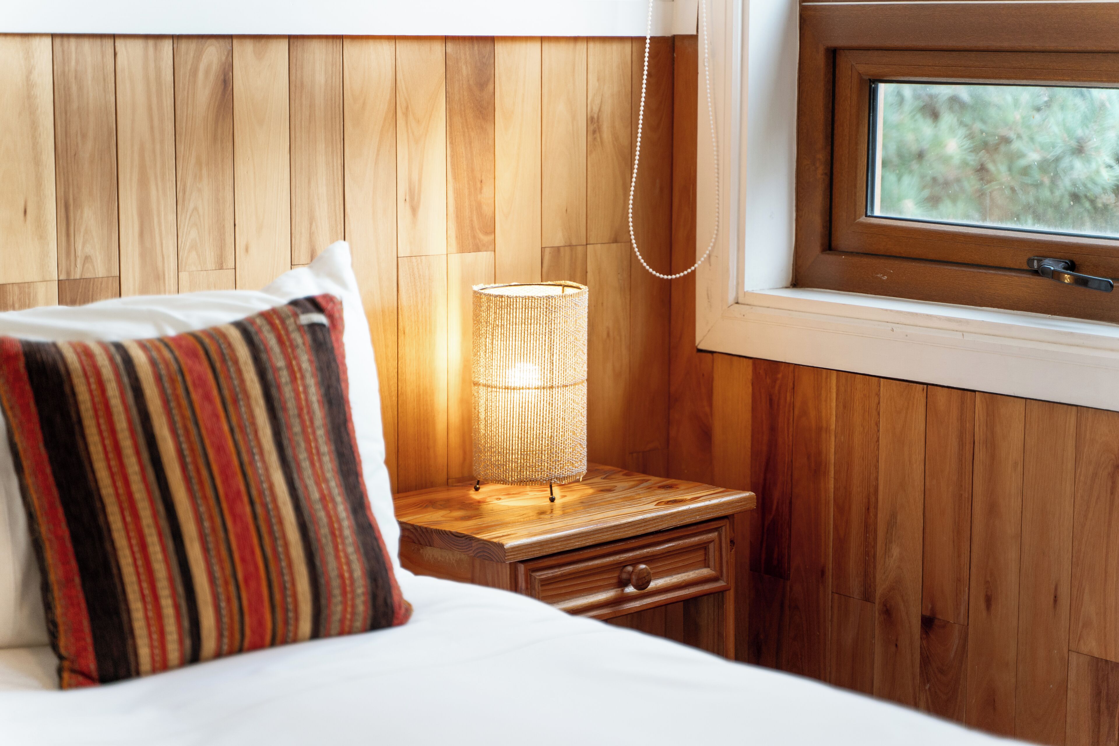 A wooden-paneled room with a bed, a bedside lamp, a nightstand, and a window.