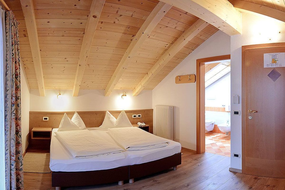 A bedroom with a wooden ceiling, a bed with white bedding, a nightstand, a wall-mounted light, and a door with a sign.