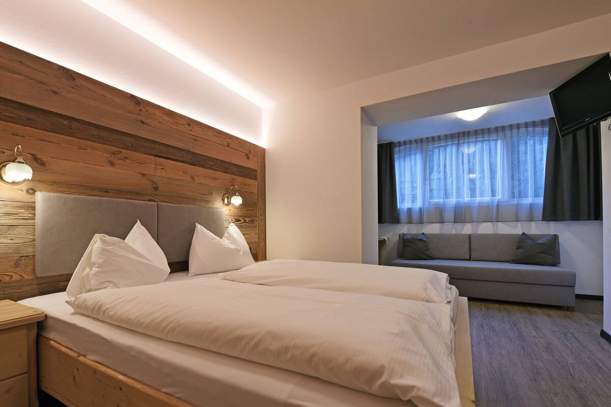 A bedroom with a wooden headboard, a bed with white bedding, a nightstand, a sofa, and a TV.