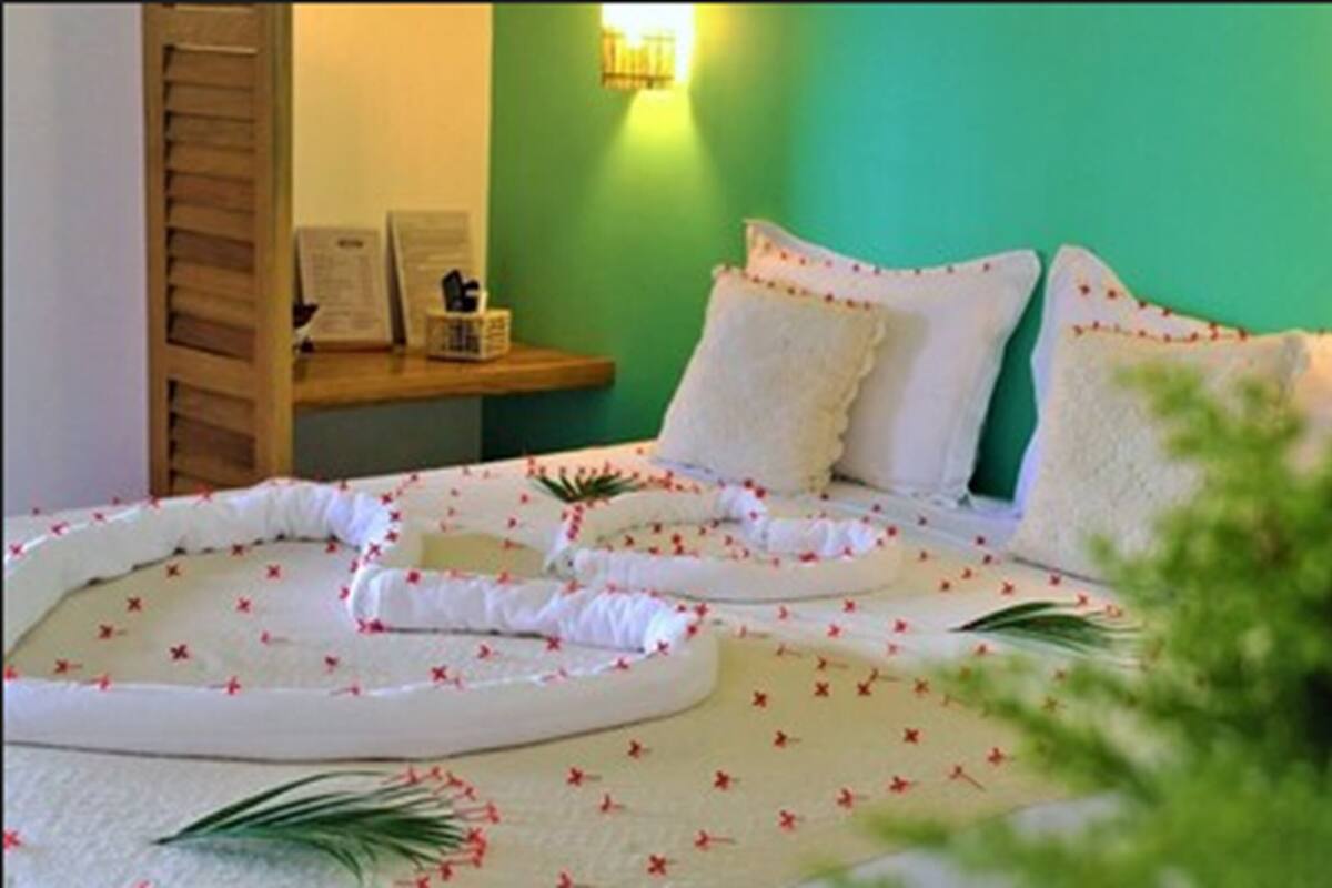 Una cama bien ordenada con sábanas blancas y almohadones decorativos, una mesita de noche con lámpara, y una pared verde con apliques.