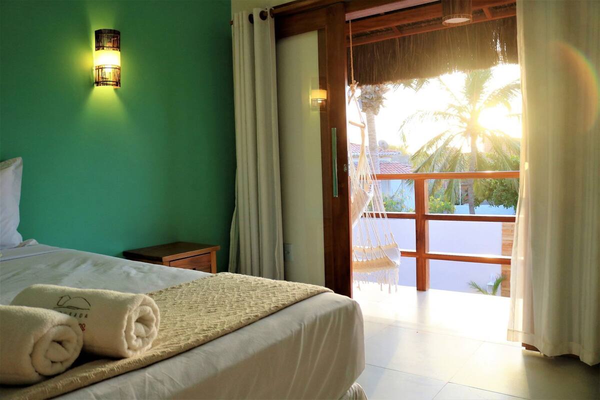 Habitación de hotel con cama, mesita de noche, hamaca y vistas a palmeras desde un balcón.