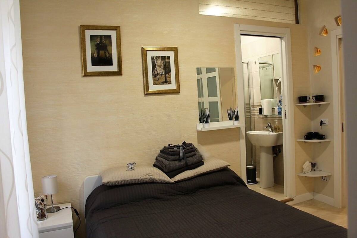 A bedroom with a bed, bedside table, wall-mounted pictures, a bathroom with a sink and mirror, and a shelf with toiletries.