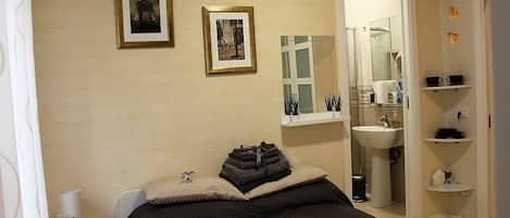 Un dormitorio con cama, mesita de noche, cuadros en la pared, baño con lavamanos y espejo, y estante con artículos de aseo.