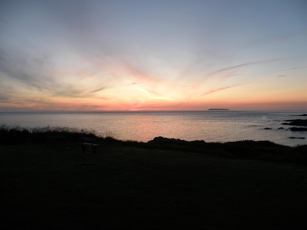 Un atardecer sobre un mar tranquilo con una isla distante, un primer plano con hierba y un banco.