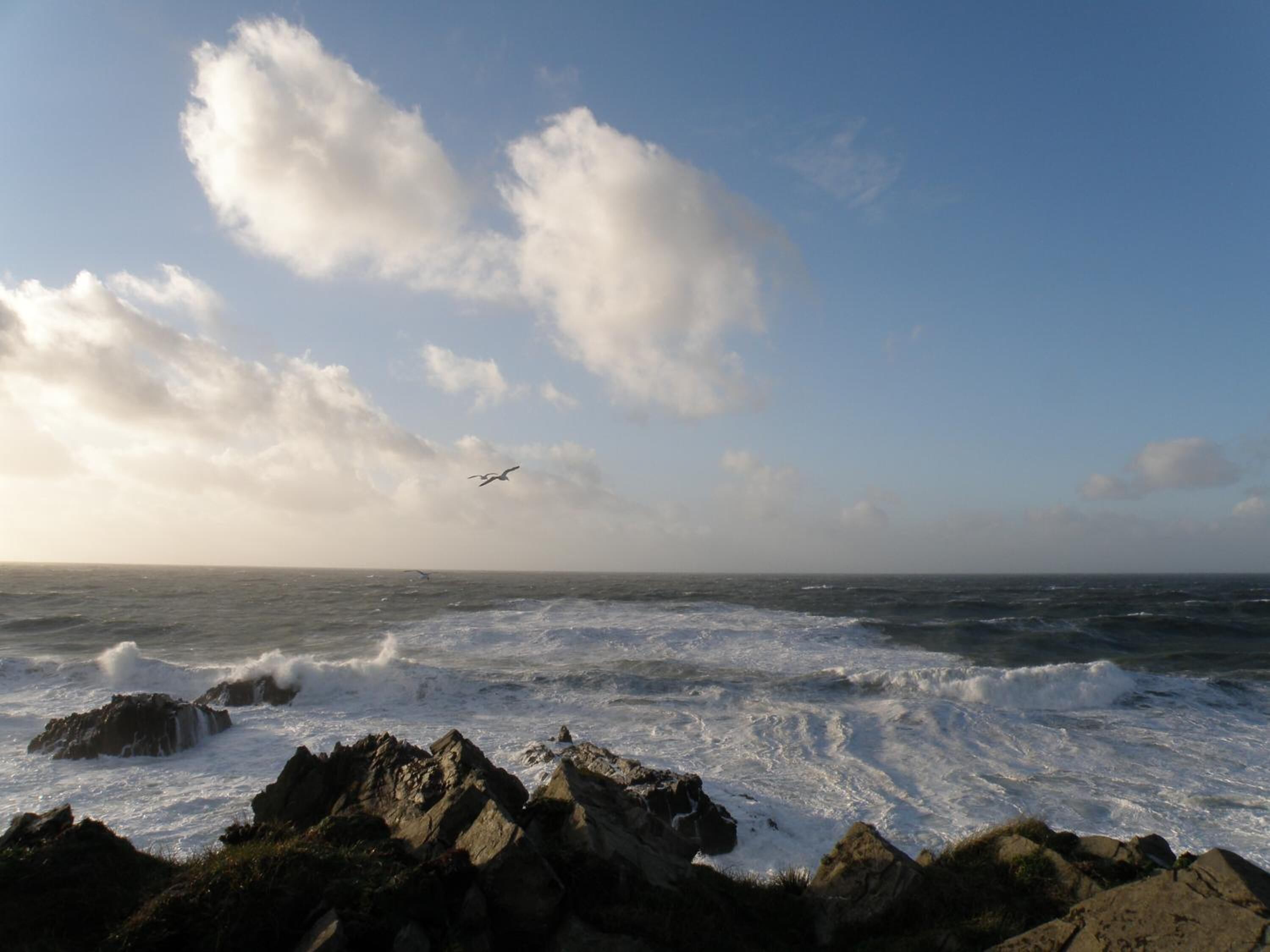 A rocky coastline with waves crashing against the rocks under a partly cloudy sky.