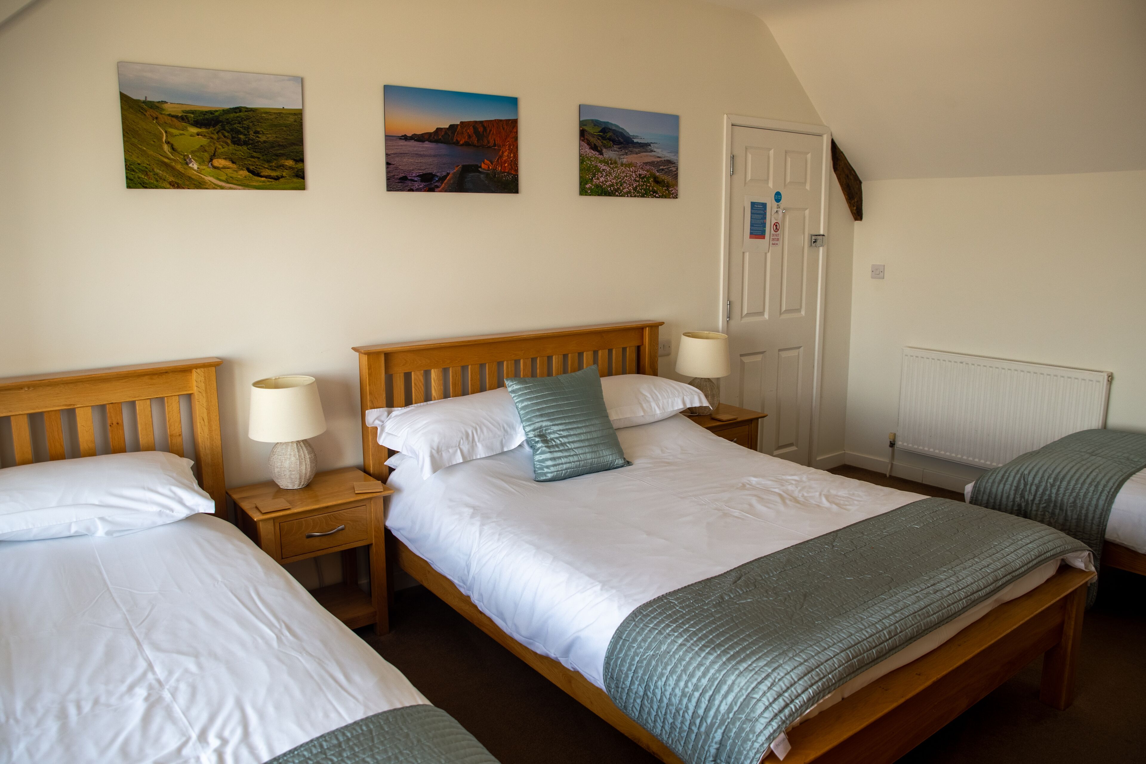 A bedroom with two single beds, a wooden headboard, bedside tables with lamps, and three landscape paintings on the wall.