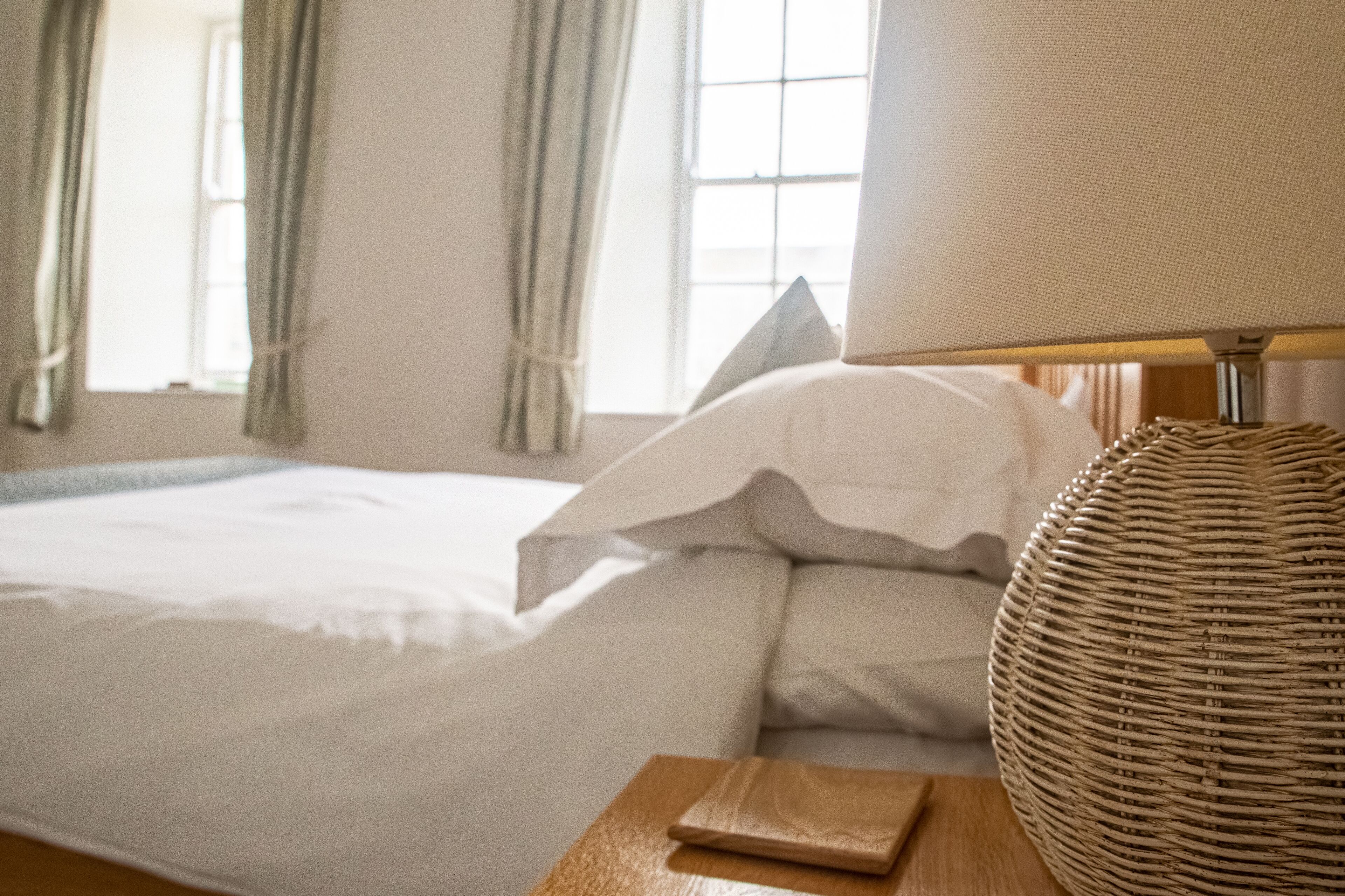 A neatly made bed with white linens, a wooden bedside table with a lamp, and a window with curtains.