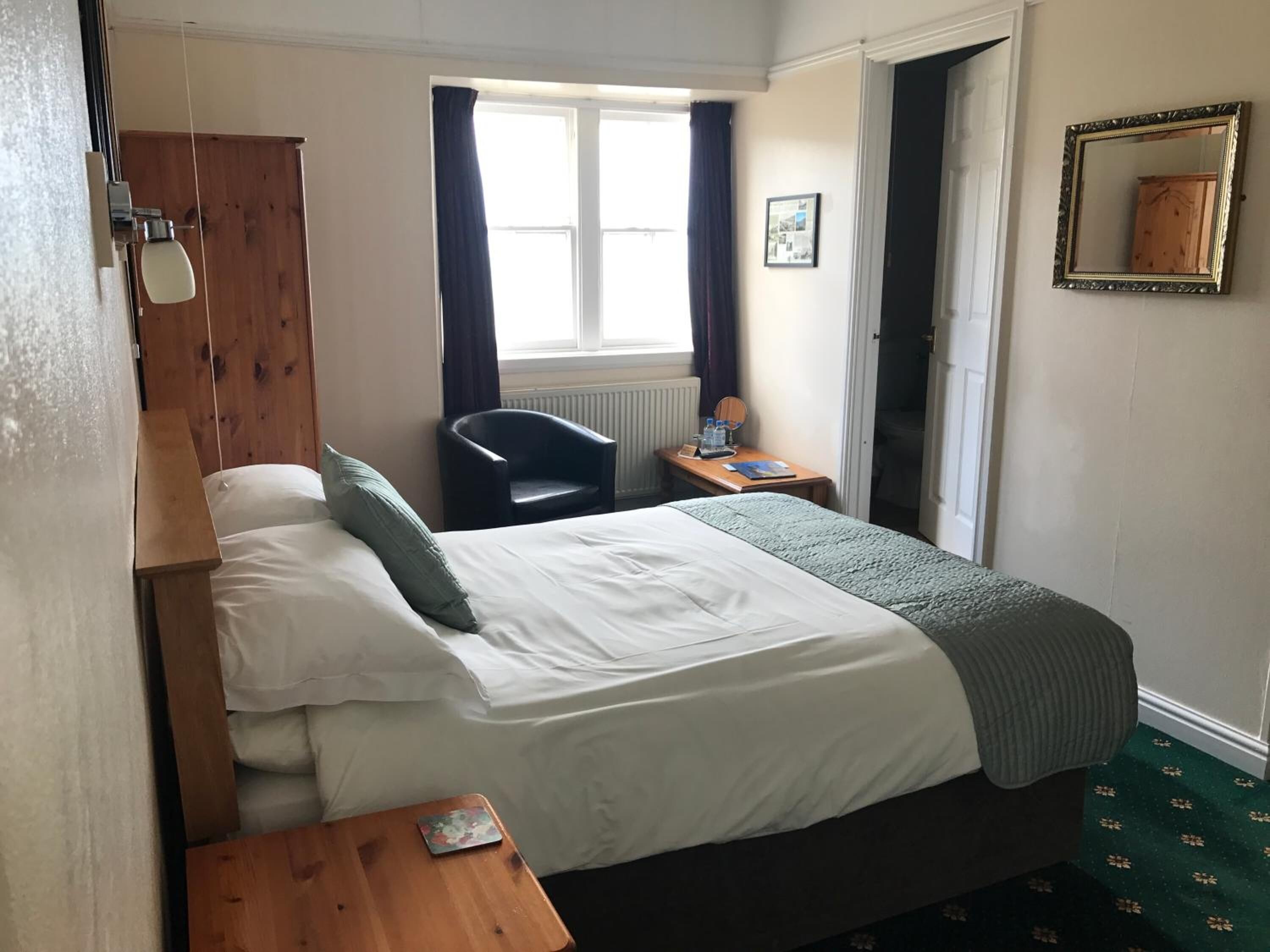Ein Hotelzimmer mit Bett, Schreibtisch, Stuhl, einem Fenster mit Vorhängen, Heizkörper und Spiegel an der Wand.