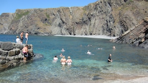 Uma praia rochosa com águas cristalinas e pessoas nadando e paradas sobre as pedras.