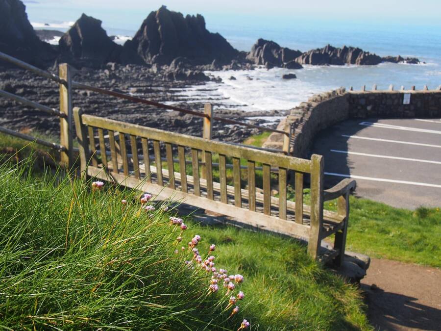 Un banc amb vista a una carretera costanera, un mur de pedra i penya-segades rocoses al fons.