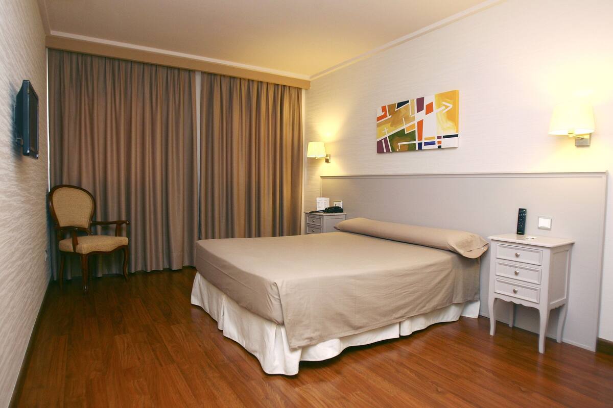 A hotel room with a bed, wooden floor, a chair, a nightstand, and a wall-mounted lamp.