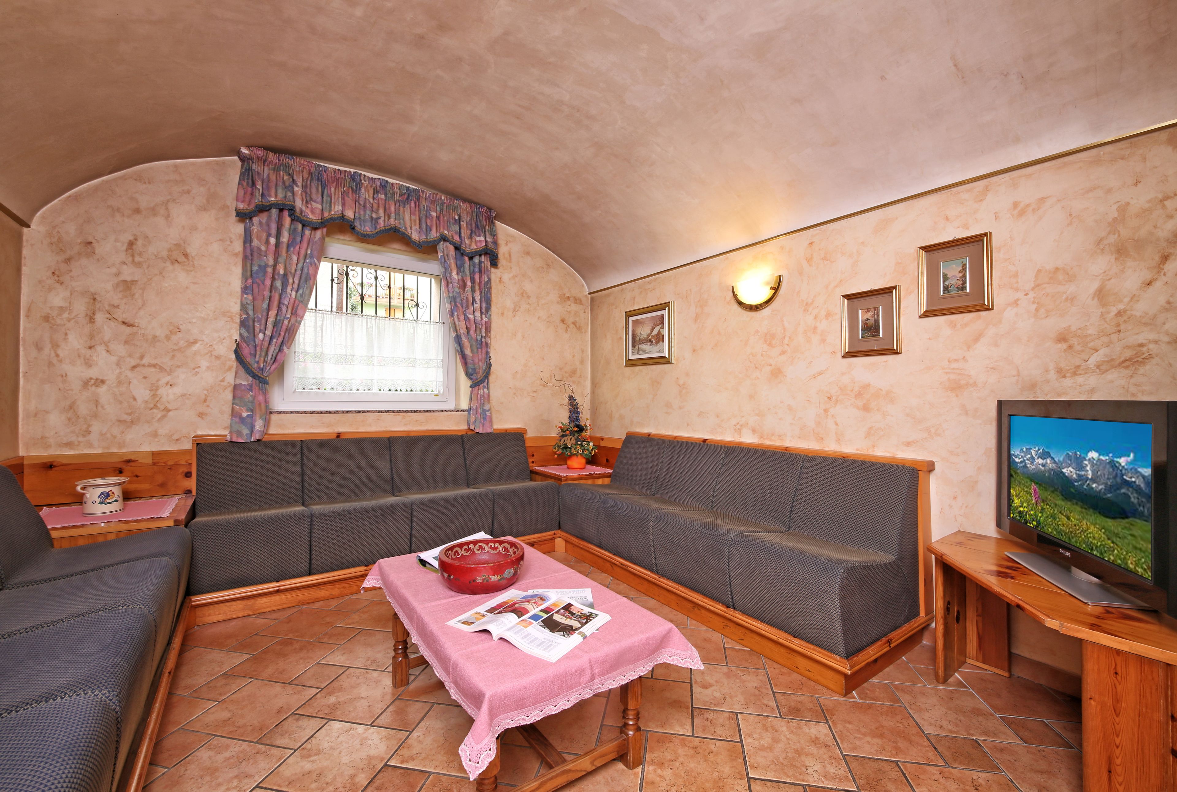 Ein gemütliches Wohnzimmer mit einer Eckcouch, einem Couchtisch aus Holz mit Zeitschriften, einem Fernseher und einem Fenster mit Vorhängen.