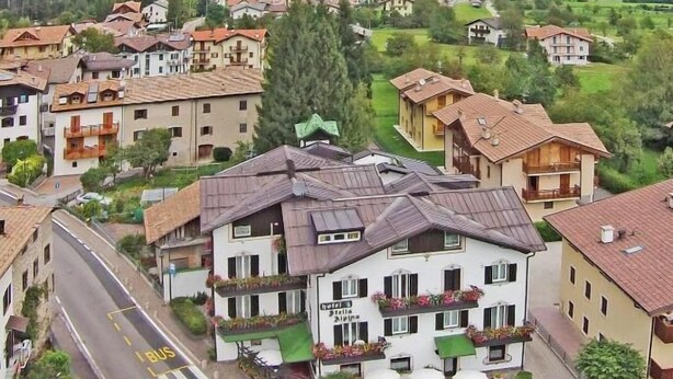 A hotel with a balcony, a road with a bus lane, and mountainous background. - Hotel Stella Alpina (Fai della Paganella)