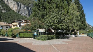 A paved parking area with a large tree in the center, buildings in the background, and a mountain. - Hotel Stella Alpina (Fai della Paganella)