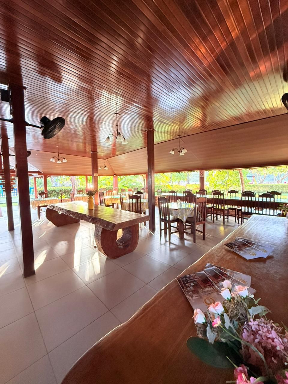 An indoor dining area with wooden tables and chairs, a ceiling with slatted design, and a view of greenery outside.