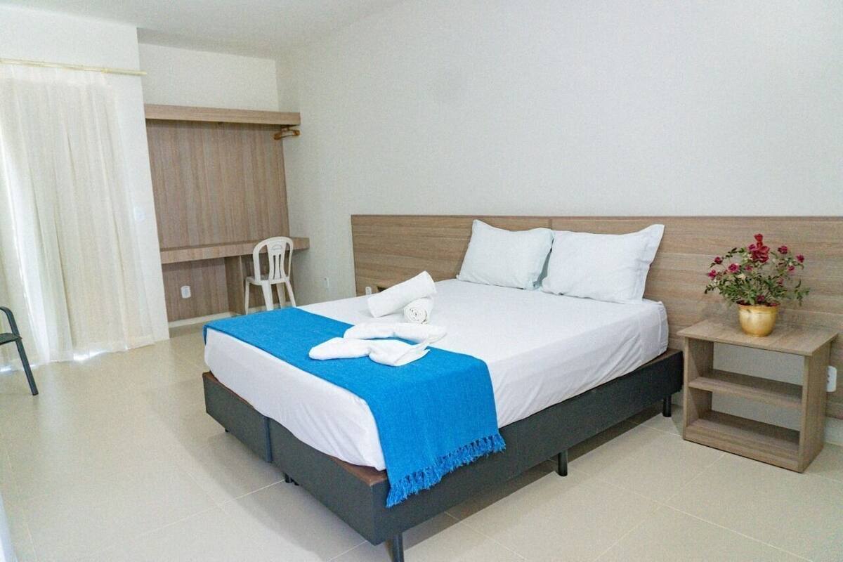 A neatly made bed with white linens and a blue throw, a wooden nightstand with a plant, and a chair in a modern room with light-colored walls and floor.