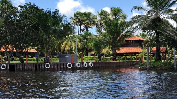 A waterfront property with a wooden fence, palm trees, and a building with a red roof. - Portal do Sol Lençóis Pousada (Barreirinhas)