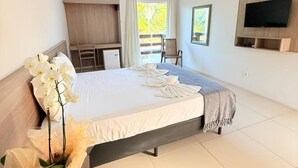 A bedroom with a bed, a TV mounted on the wall, a desk with a remote control, and a window with curtains. - Portal do Sol Lençóis Pousada (Barreirinhas)