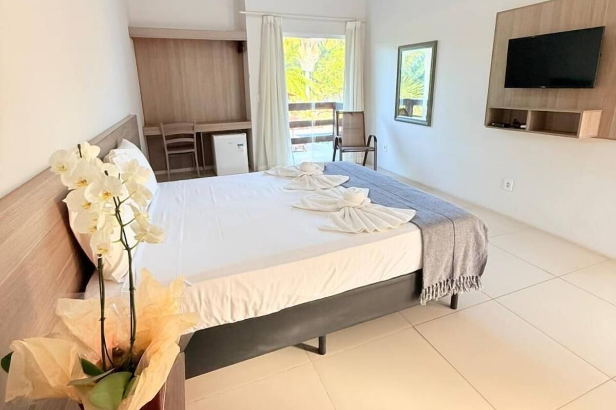 A bedroom with a bed, a TV mounted on the wall, a desk with a remote control, and a window with curtains.