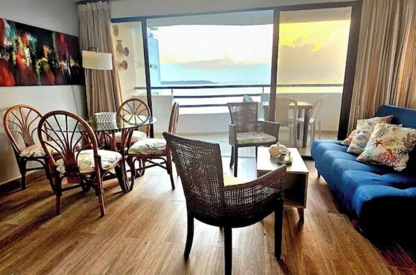 A room with a wooden floor, a blue sofa with floral cushions, wicker chairs, a small table, and a large window with a view of the sea. - Edificio Marina del Rey (Cartagena)