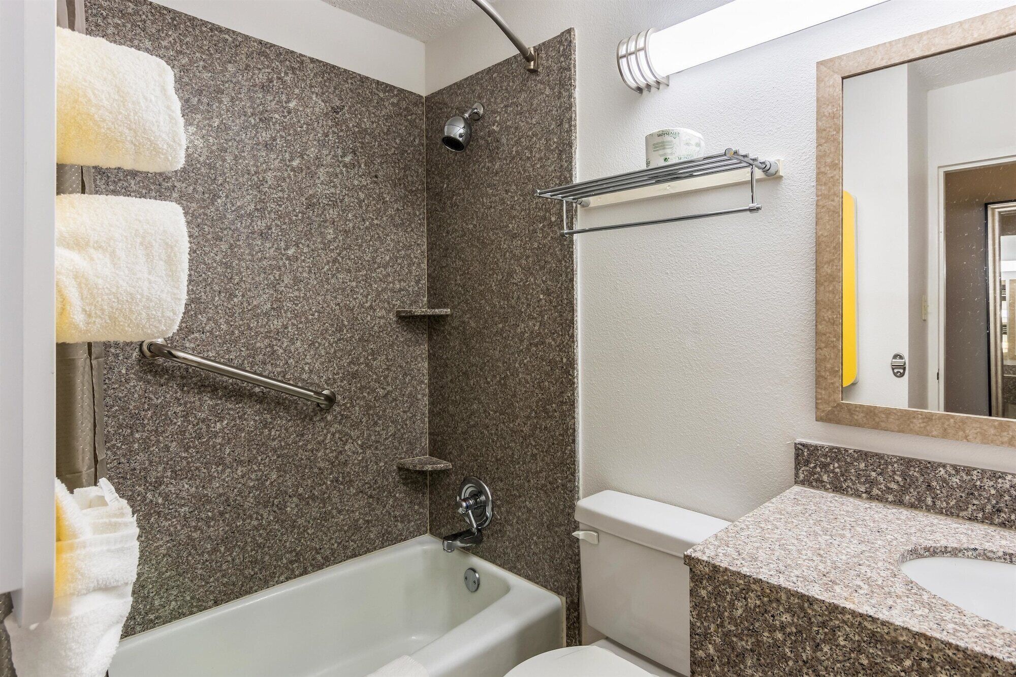Un baño con bañera, inodoro y ducha. Toallas cuelgan de un perchero, y hay un estante con artículos de aseo.