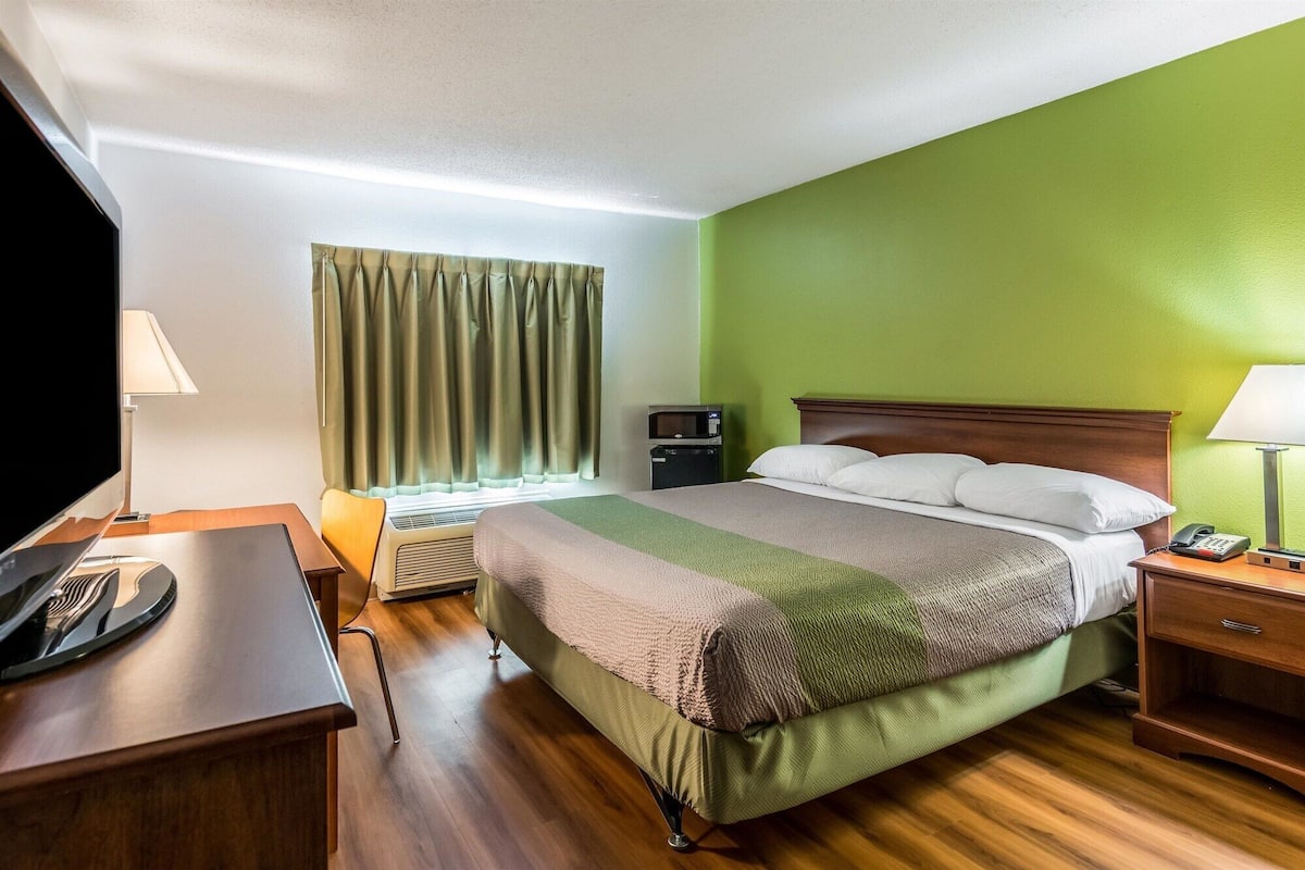 Habitación de hotel con cama, escritorio con televisor, mesita de noche con lámpara, calefactor y ventana con cortinas.