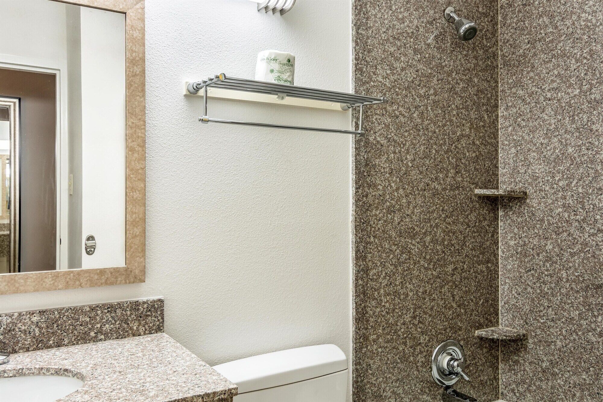 A bathroom with a granite countertop, a toilet, a shower with a glass enclosure, and a towel rack with a bottle of hand soap.